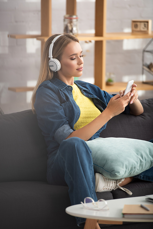 Young blonde woman in headphones texting on smartphone while sitting on couch on blurred background