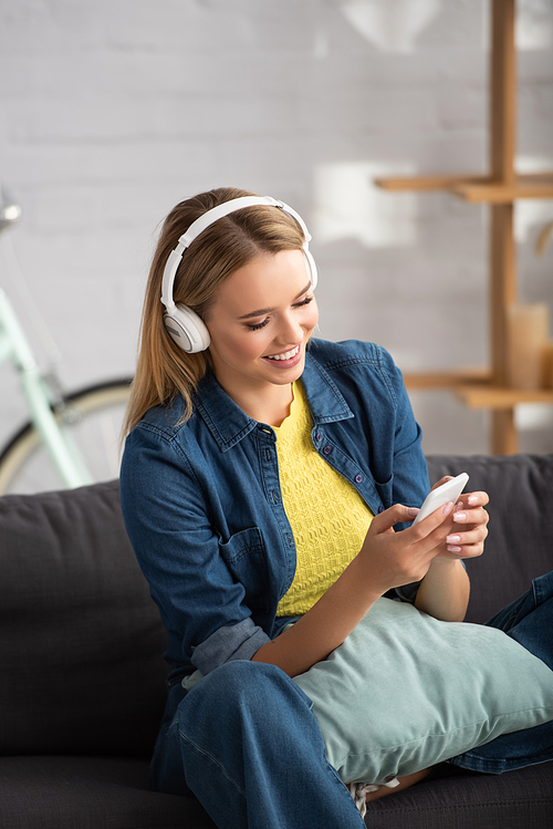 Smiling blonde woman in headphones texting on smartphone at home on blurred background