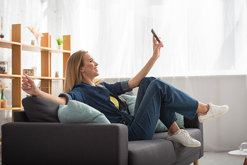 Full length of smiling blonde woman with smartphone taking selfie while sitting on couch on blurred background