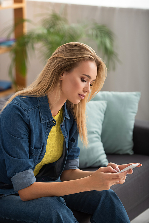 Young blonde woman texting on smartphone at home on blurred background