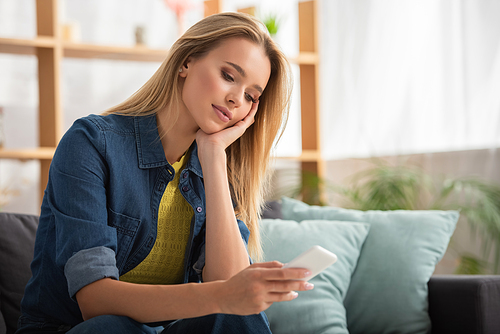 Young blonde woman looking at smartphone while sitting on couch at home on blurred background