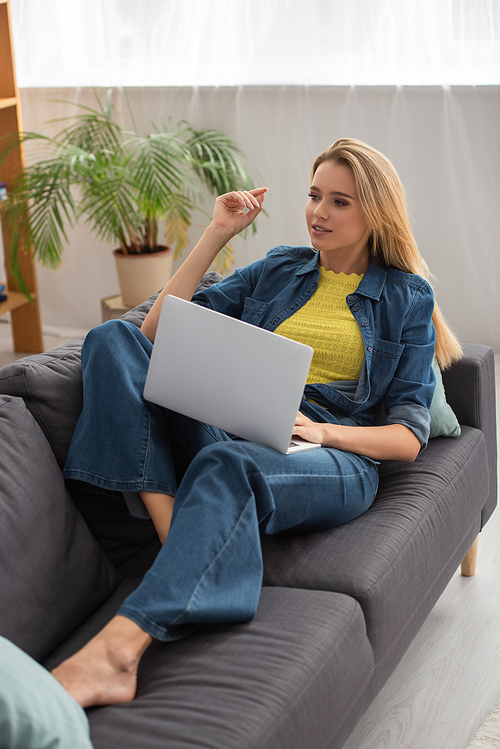 Full length of young woman looking away while using laptop on couch at home