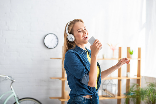 Cheerful blonde woman in headphones singing while holding smartphone at home on blurred background
