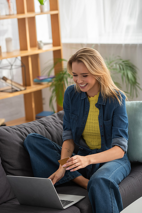 Happy blonde woman with credit card laughing while sitting on couch near laptop on blurred background