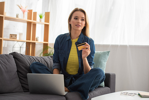 Young blonde woman with credit card looking away while sitting near laptop on couch on blurred background