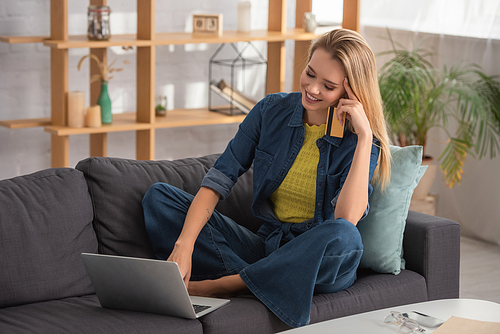 Cheerful blonde woman with credit card using laptop while sitting on couch at home on blurred background
