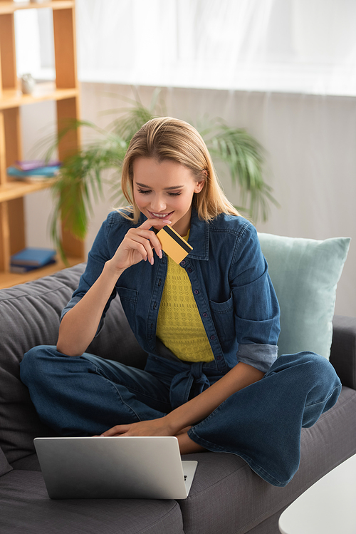 Smiling young blonde woman with credit card looking at laptop while sitting on couch on blurred background