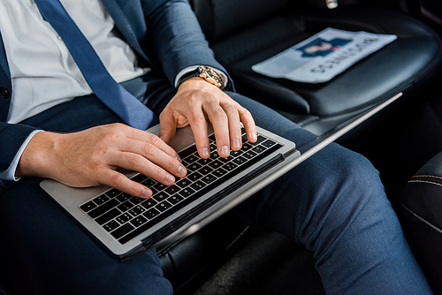Cropped view of man in formal wear using laptop near newspaper on blurred background in car