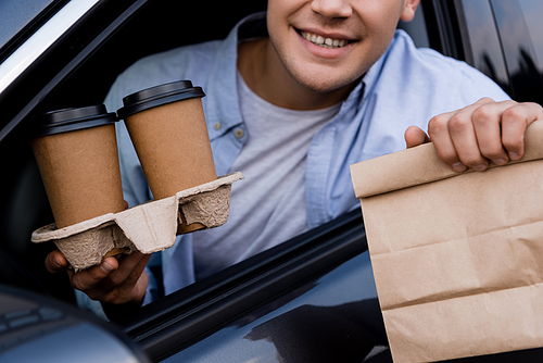cropped view of smiling man holding paper bag and coffee to go in car, blurred foreground
