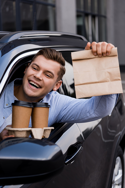happy man holding disposable cup and paper bag while sitting in car