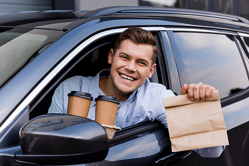 cheerful driver holding coffee to go and paper bag while smiling at camera