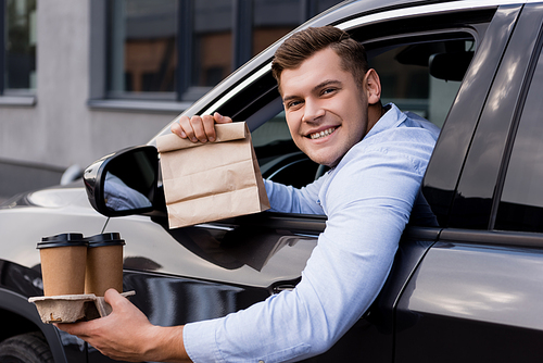 happy man holding disposable cups and paper bag while sitting in car and 
