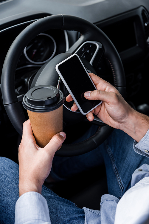 cropped view of man holding coffee to go and smartphone with blank screen near steering wheel