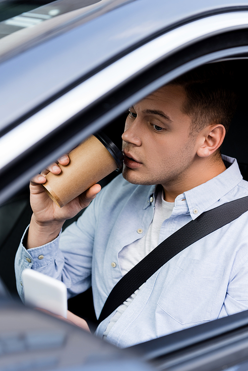 man messaging on mobile phone and drinking coffee to go in car on blurred foreground