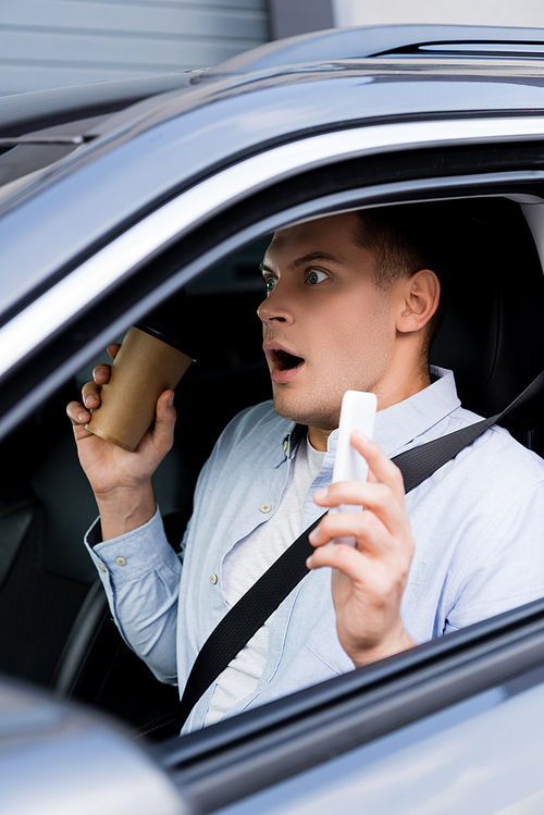 shocked man holding coffee to go and mobile phone at drivers seat in car, blurred foreground