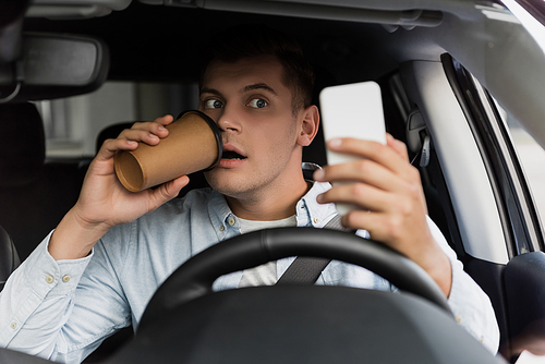 surprised man drinking coffee to go and using smartphone at steering wheel in car, blurred foreground