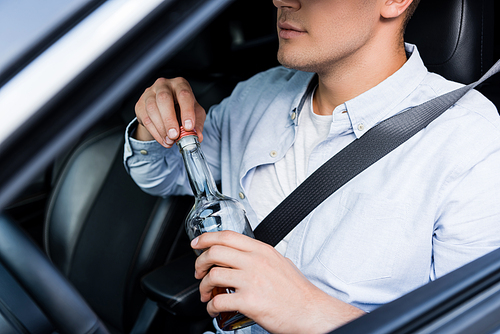 cropped view of man opening bottle of alcohol while sitting at drivers seat in car, blurred foreground