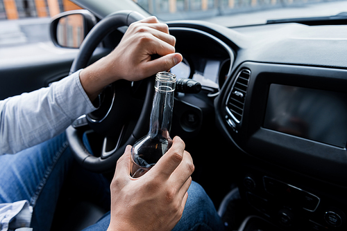 partial view of man holding bottle of whiskey while driving car