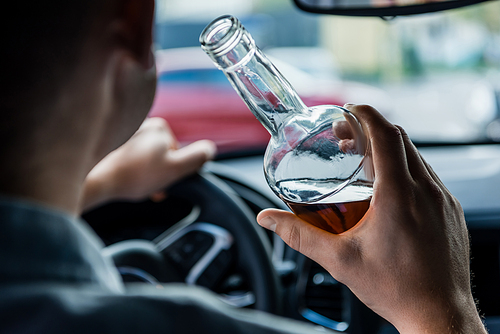 cropped view of man holding bottle of alcohol while driving car, blurred foreground