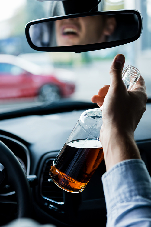 partial view of drunk man driving car and holding bottle of whiskey, blurred foreground