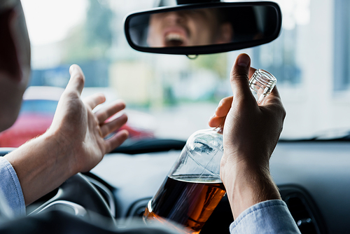 partial view of angry drunk man with bottle of whiskey shouting and gesturing while driving car, blurred foreground