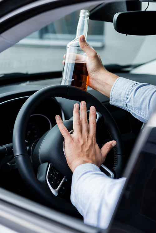 partial view of man beeping while driving car and holding bottle of whiskey, blurred foreground