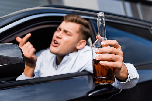 drunk man with bottle of alcohol showing come here gesture while looking out car window, blurred foreground