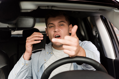 drunk man holding flask with alcohol and showing middle finger in car, blurred foreground