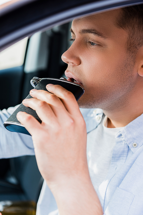 young man drinking alcohol from flask while driving car, blurred foreground