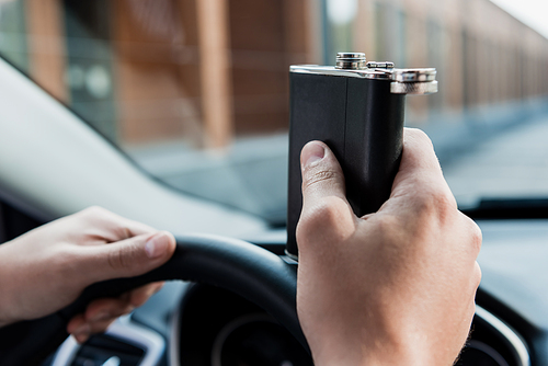 cropped view of man holding flask with alcohol while driving car, blurred background