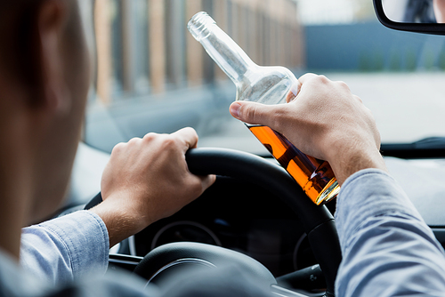 partial view of man driving car while holding bottle of whiskey, blurred foreground