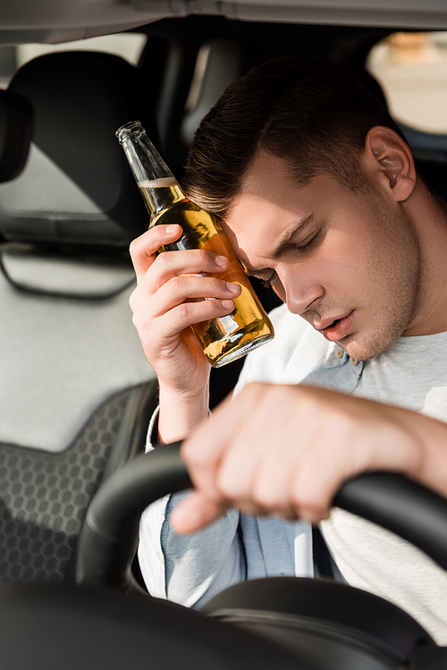 drunk man holding bottle of alcohol near head while sitting at steering wheel in car, blurred foreground