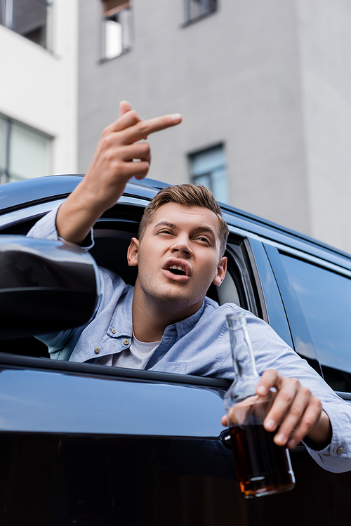 drunk, aggressive man with bottle of whiskey showing middle finger while looking out car window, blurred foreground