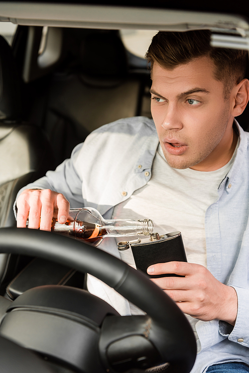 drunk man pouring whiskey from bottle to flask while sitting in car, blurred foreground