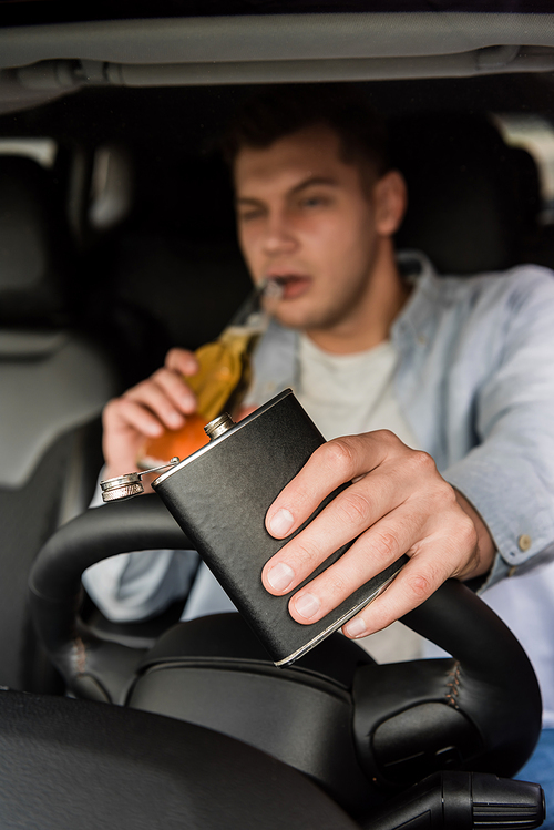 young man holding flask and drinking whiskey while driving car, blurred background