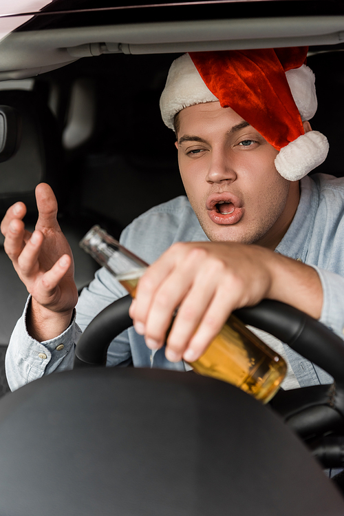 drunk man in santa hat holding bottle of alcohol and gesturing while driving car on blurred foreground