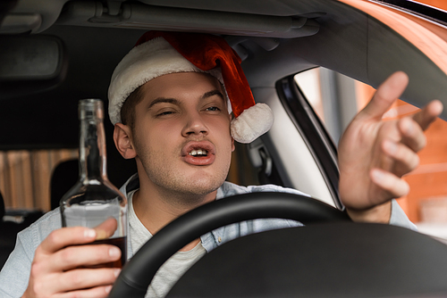 displeased, drunk man in santa hat gesturing while sitting at steering wheel with bottle of whiskey, blurred foreground
