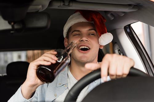 drunk, excited man in santa hat driving car and holding bottle of whiskey on blurred foreground