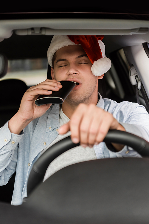 drunk man in santa hat drinking from flask with closed eyes while driving car on blurred foreground