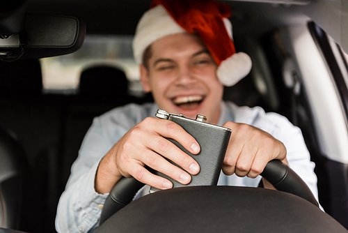 laughing, drunk man in santa hat holding flask with alcohol while driving car on blurred background