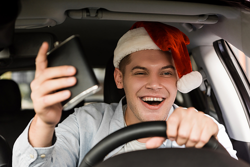 drunk, cheerful man in santa hat holding flask with alcohol while driving car, blurred foreground