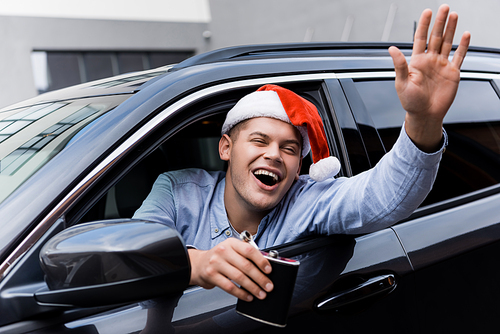 drunk, excited man in santa hat waving hand and holding flask of alcohol while looking out car window