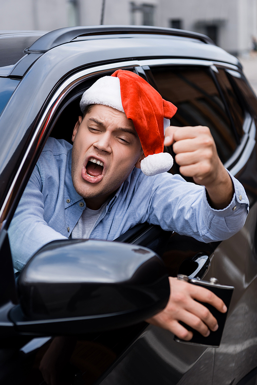 aggressive, drunk man in santa hat, with flask of alcohol, showing clenched fist while looking out car window, blurred foreground