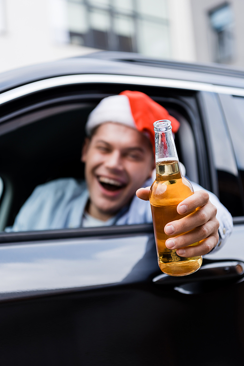 drunk, laughing man in santa hat holding bottle of whiskey in outstretched hand while sitting in car, blurred foreground