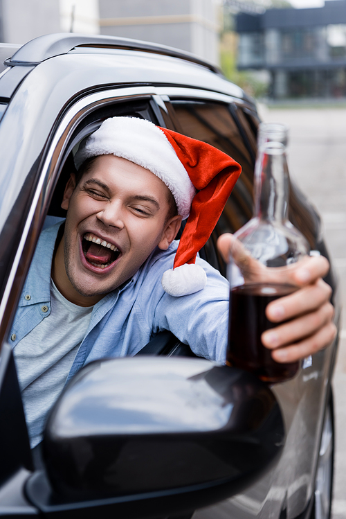 drunk man in santa hat grimacing and holding bottle of whiskey in car on blurred foreground