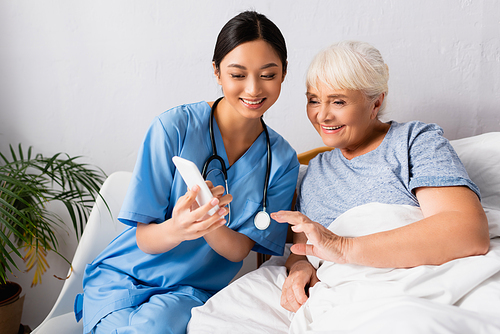 cheerful asian nurse using mobile phone near happy senior woman lying in bed