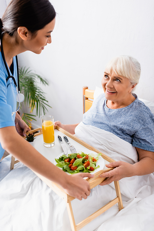 smiling asian nurse holding tray with breakfast near happy elderly woman in bed