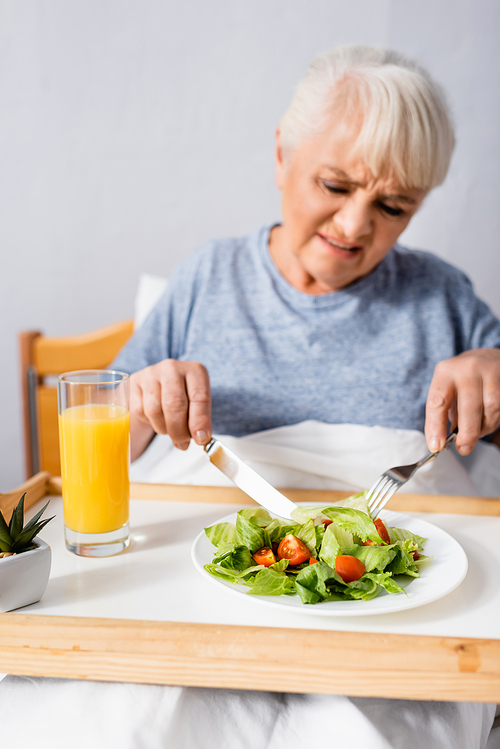 displeased aged woman having breakfast in hospital, blurred foreground