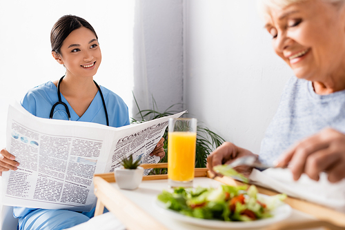 smiling asian nurse holding newspaper near pleased senior woman having breakfast on blurred foreground