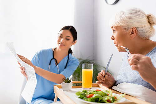 amazed senior woman having breakfast and looking at smiling asian nurse reading newspaper on blurred background